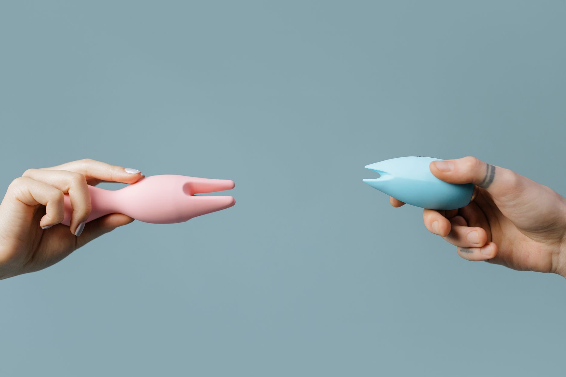 Two hands holding colorful silicone vibrators together against a pastel background