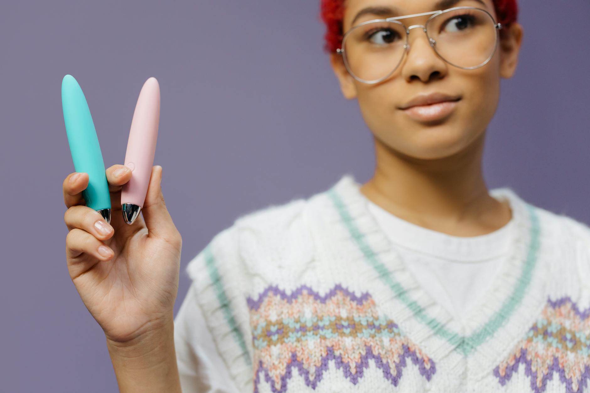 A person holding two colorful vibrators against a purple background, exploring options for pleasure.